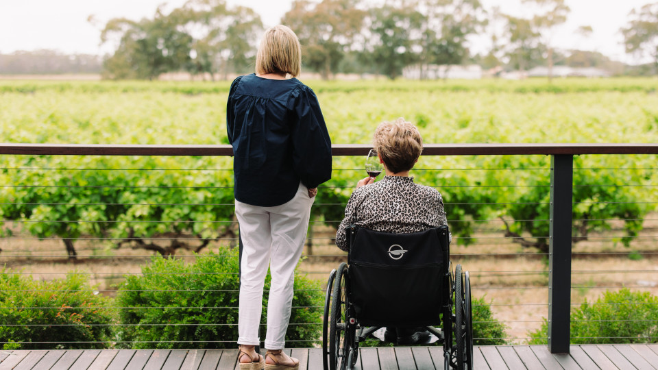 A woman standing on the left and another sitting in a wheelchair to the right. Both are on a wooden deck facing away looking out at vineyards.