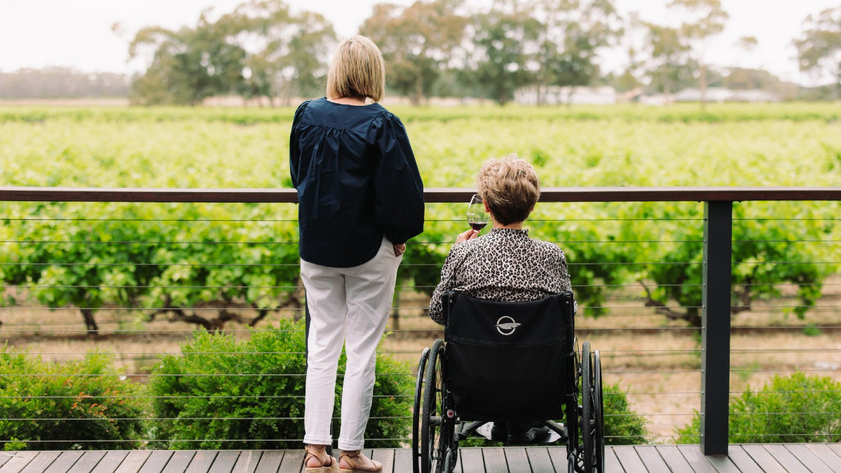 Two women, one in a wheelchair, on a deck with backs to the camera, looking out at vineyards.